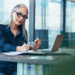 A woman sitting at a table with a laptop and writing on a piece of paper about definition, types, benefits, and low barrier details regarding housing loans