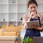 A woman standing in front of boxes while looking for cash advance, merchant, and installment advances with the lowest rates using her tablet